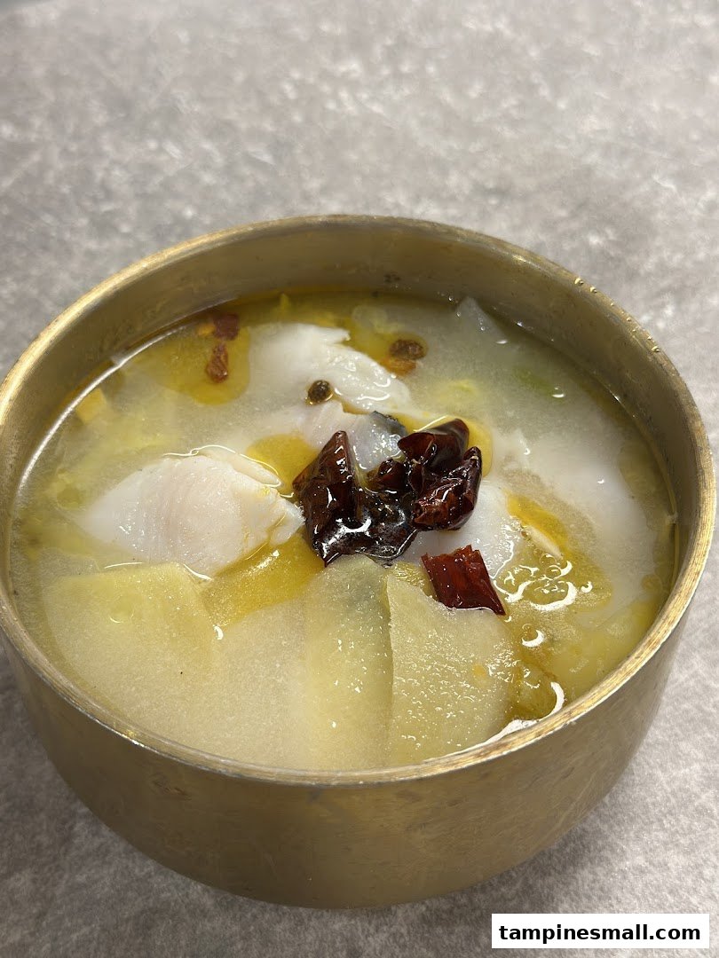 A close-up of a bowl of fish soup with vegetables and chili in a brass bowl.