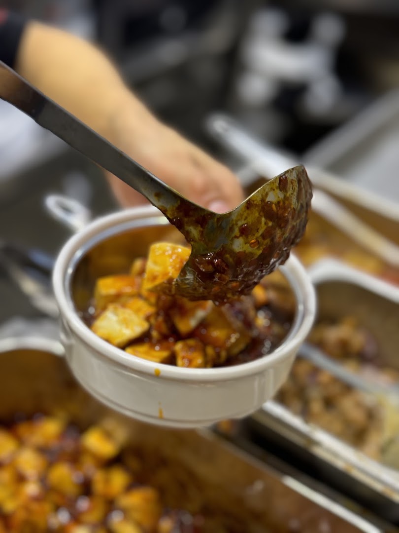 A ladle serves a spicy, saucy dish, likely Mapo Tofu, into a small bowl.
