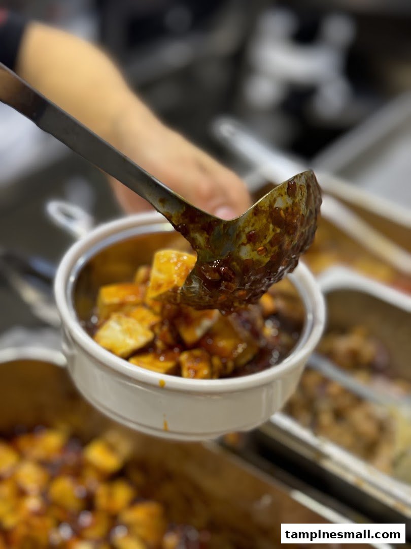 A ladle serves a spicy, saucy dish, likely Mapo Tofu, into a small bowl.