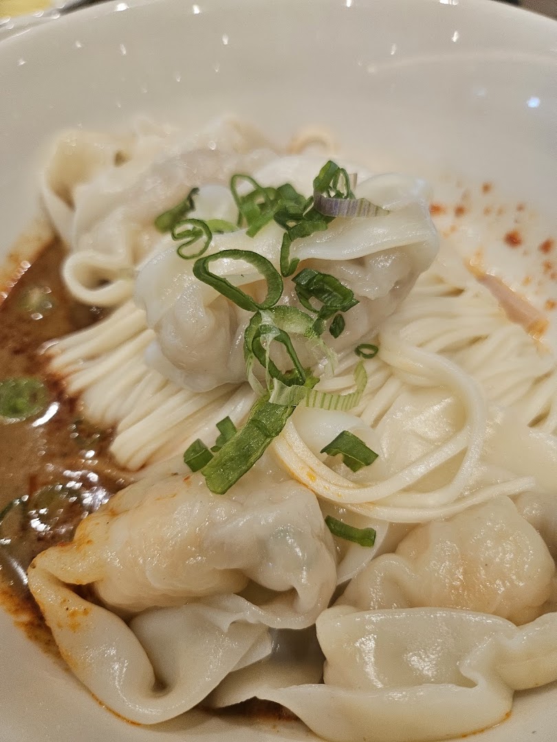 A close-up of a bowl of noodles with dumplings, chili oil, and chopped spring onions.