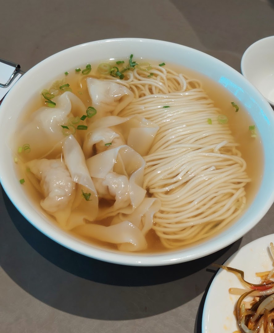 A close-up of a bowl of clear noodle soup with dumplings and chopped green onions.