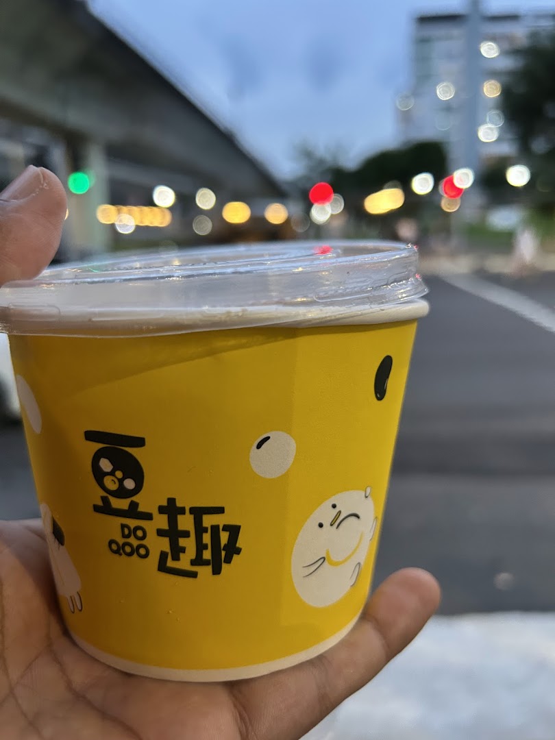 A hand holds a yellow cup from Do Qoo 豆趣 with cute characters, against a blurred urban background.