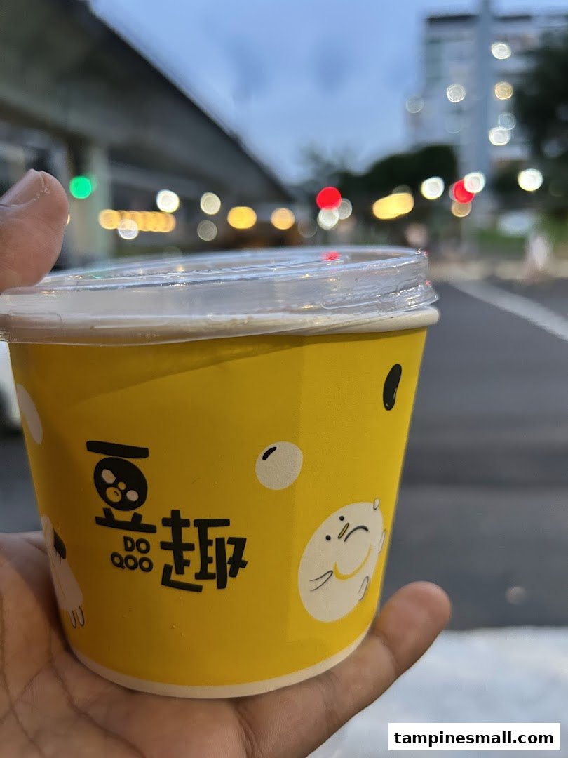A hand holds a yellow cup from Do Qoo 豆趣 with cute characters, against a blurred urban background.