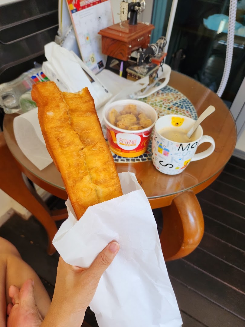 A hand holds a golden-brown fried dough stick (youtiao) with a drink and snacks on a table.