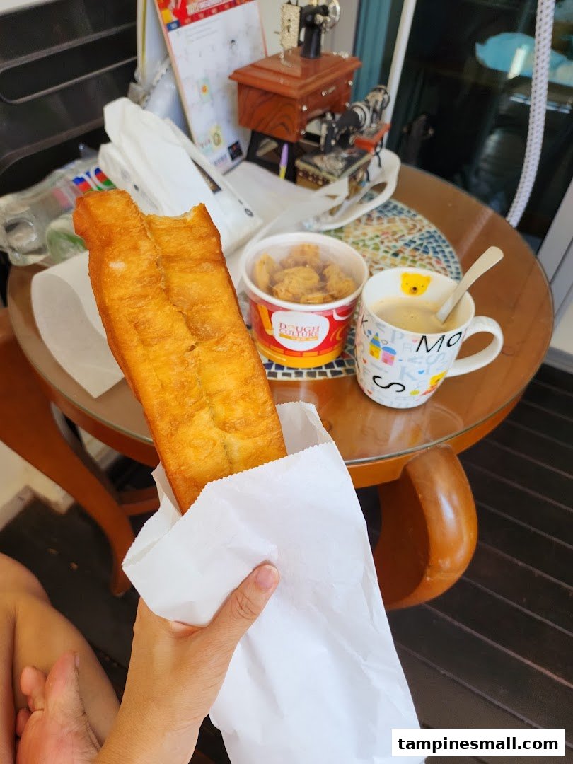 A hand holds a golden-brown fried dough stick (youtiao) with a drink and snacks on a table.