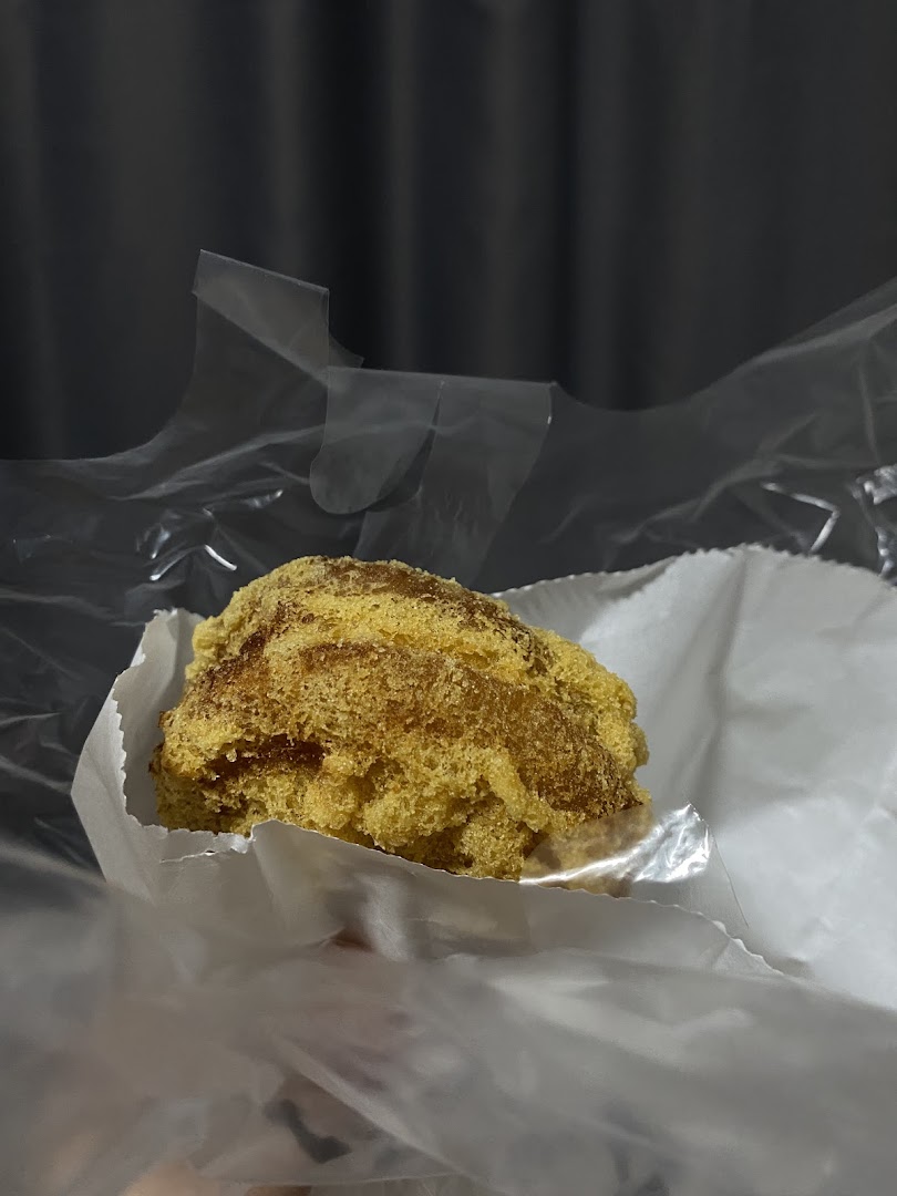 A close-up of a golden-brown fried dough pastry, possibly a floss bun, in paper and plastic packaging.