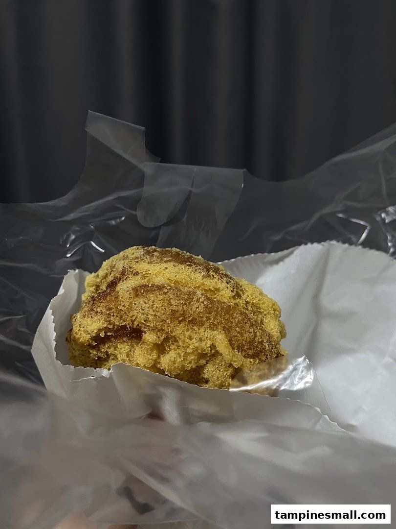 A close-up of a golden-brown fried dough pastry, possibly a floss bun, in paper and plastic packaging.