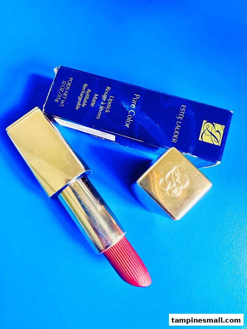 An Estée Lauder Pure Color lipstick in a gold case with its blue product box on a blue background.