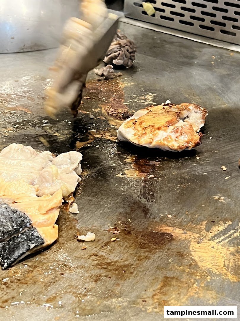 Close-up of chicken and salmon cooking on a sizzling teppanyaki grill with a spatula.
