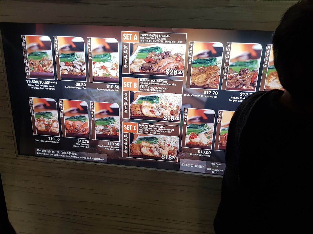 A customer views a brightly lit digital menu board displaying various teppan-yaki dishes and prices.