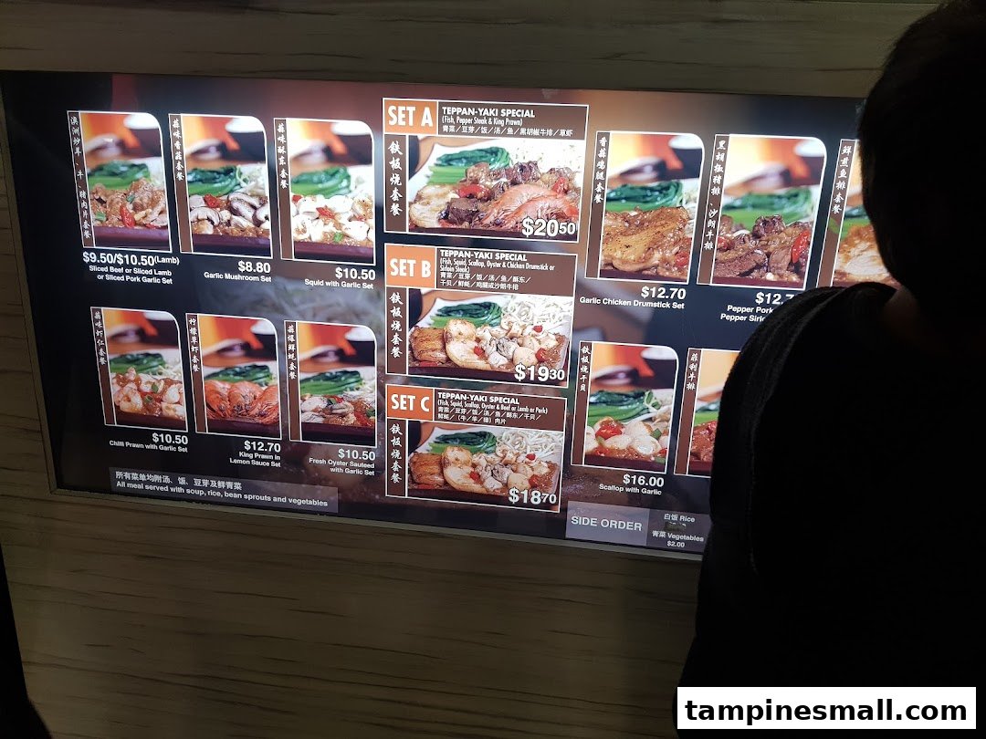 A customer views a brightly lit digital menu board displaying various teppan-yaki dishes and prices.
