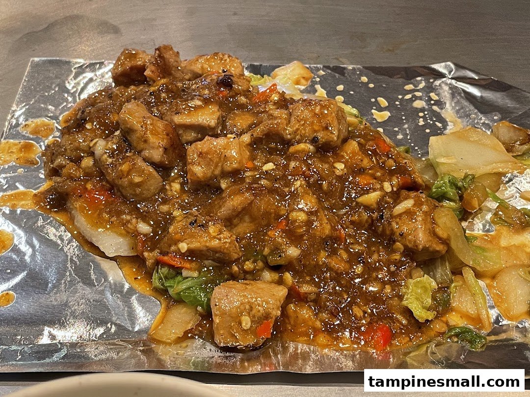 A close-up of a savory teppanyaki dish featuring saucy meat cubes and vegetables on foil.