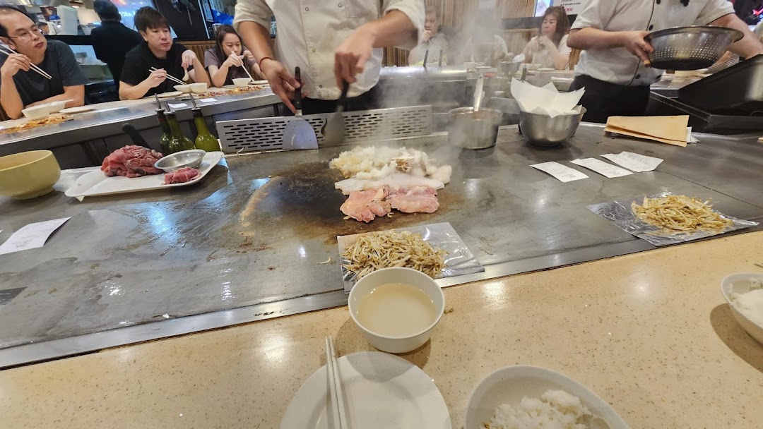A chef prepares fresh ingredients on a sizzling teppanyaki grill for seated customers.