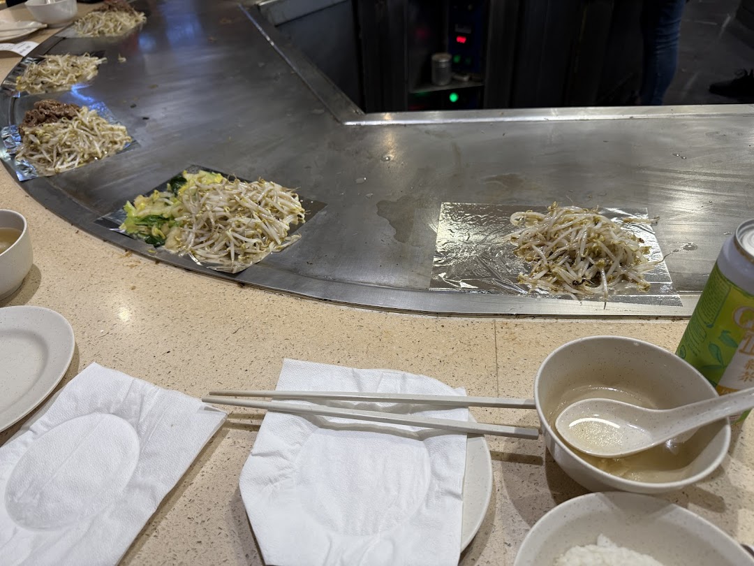 A close-up of a teppan-yaki grill with bean sprouts and vegetables cooking, alongside customer place settings.