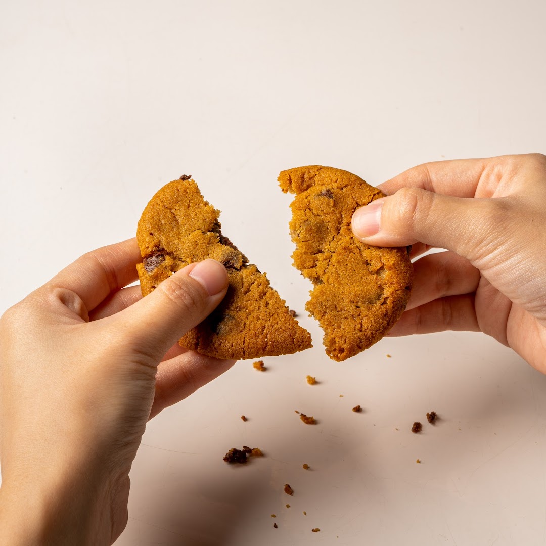 Hands breaking a fresh Famous Amos chocolate chip cookie, revealing its soft, chewy interior.