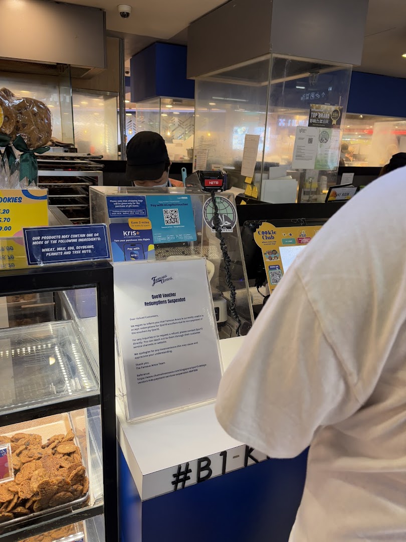 A bustling Famous Amos counter with a staff member, fresh cookies on display, and various payment signs.