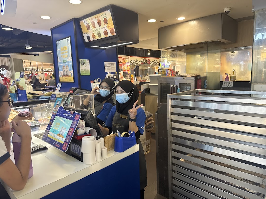 Two staff members at the Famous Amos counter serving a customer amidst freshly baked cookie displays.