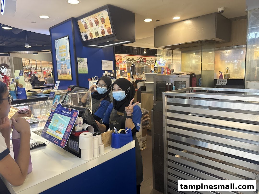 Two staff members at the Famous Amos counter serving a customer amidst freshly baked cookie displays.