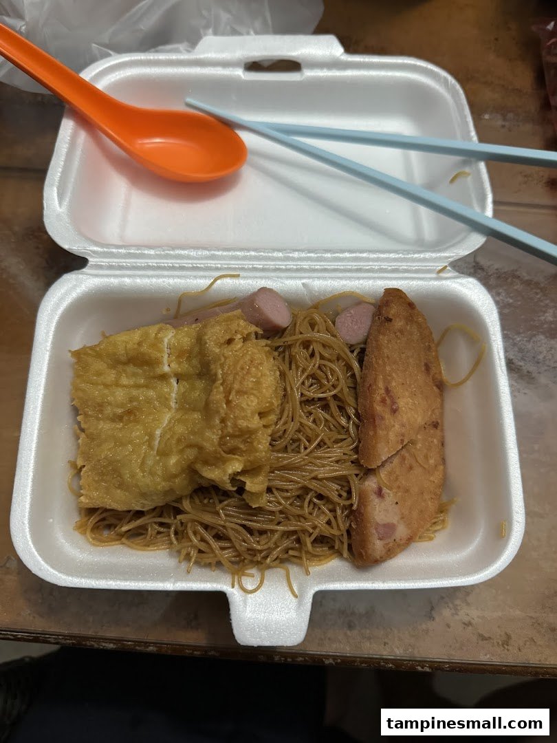 A takeaway box filled with noodles, omelette, fishcake, and sausages, with utensils.
