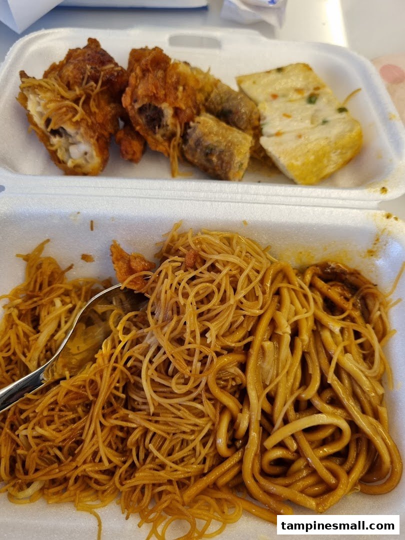 A takeaway container featuring fried noodles, crispy chicken wings, spring rolls, and fishcake.