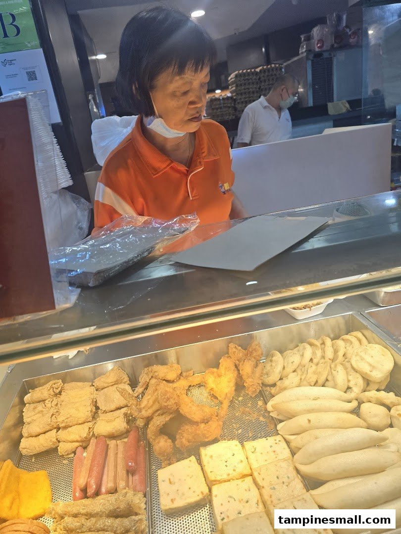 A staff member serves customers behind a counter displaying various fried and processed food items.