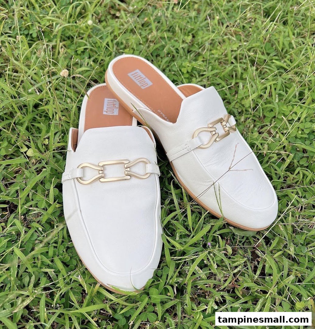 A pair of stylish white FitFlop mules with gold chain detail resting on green grass.