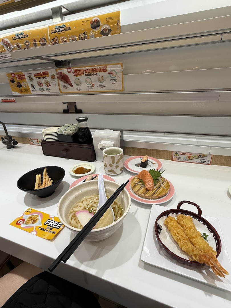 Various sushi and Japanese dishes, including udon and tempura, on a table next to a conveyor belt.