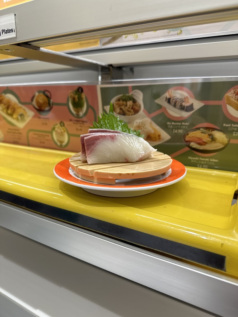 A fresh sushi dish on a conveyor belt with menu items and prices displayed in the background.