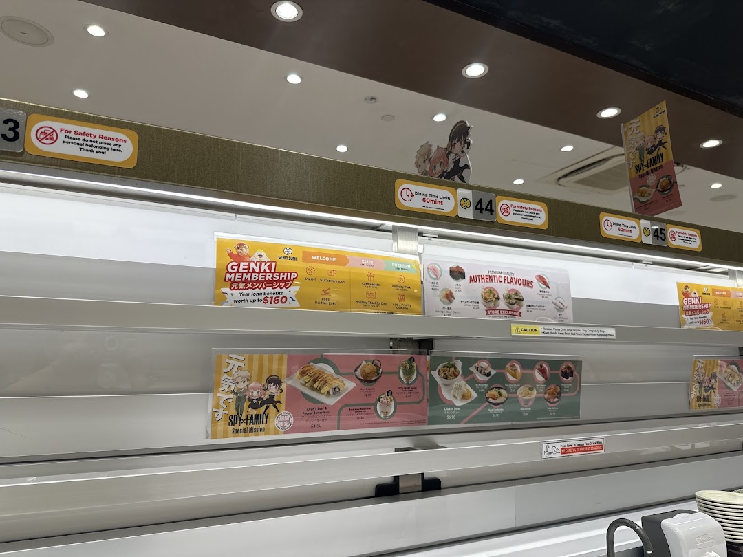 Interior view of Genki Sushi, showcasing its conveyor belt system with various promotional signs and food displays.