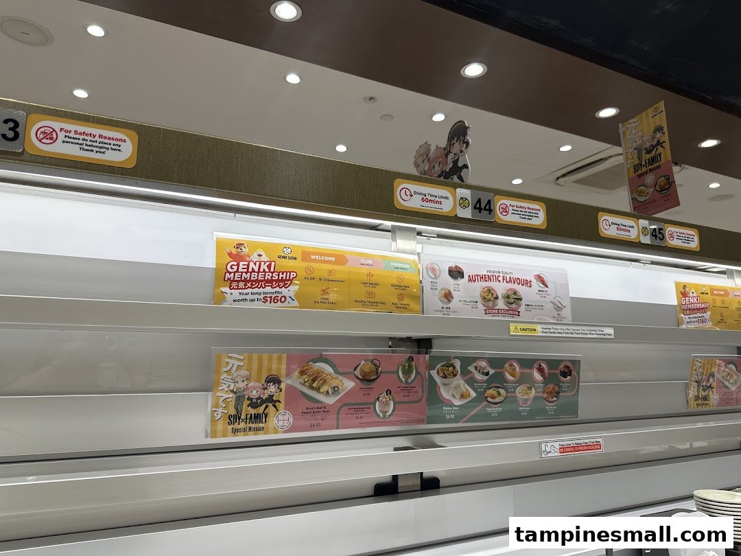 Interior view of Genki Sushi, showcasing its conveyor belt system with various promotional signs and food displays.