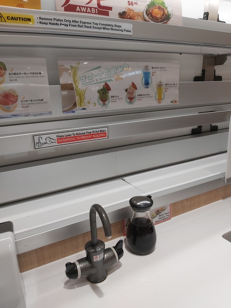 A close-up of a Genki Sushi dining counter with menu displays for desserts, mini-dons, and safety instructions.