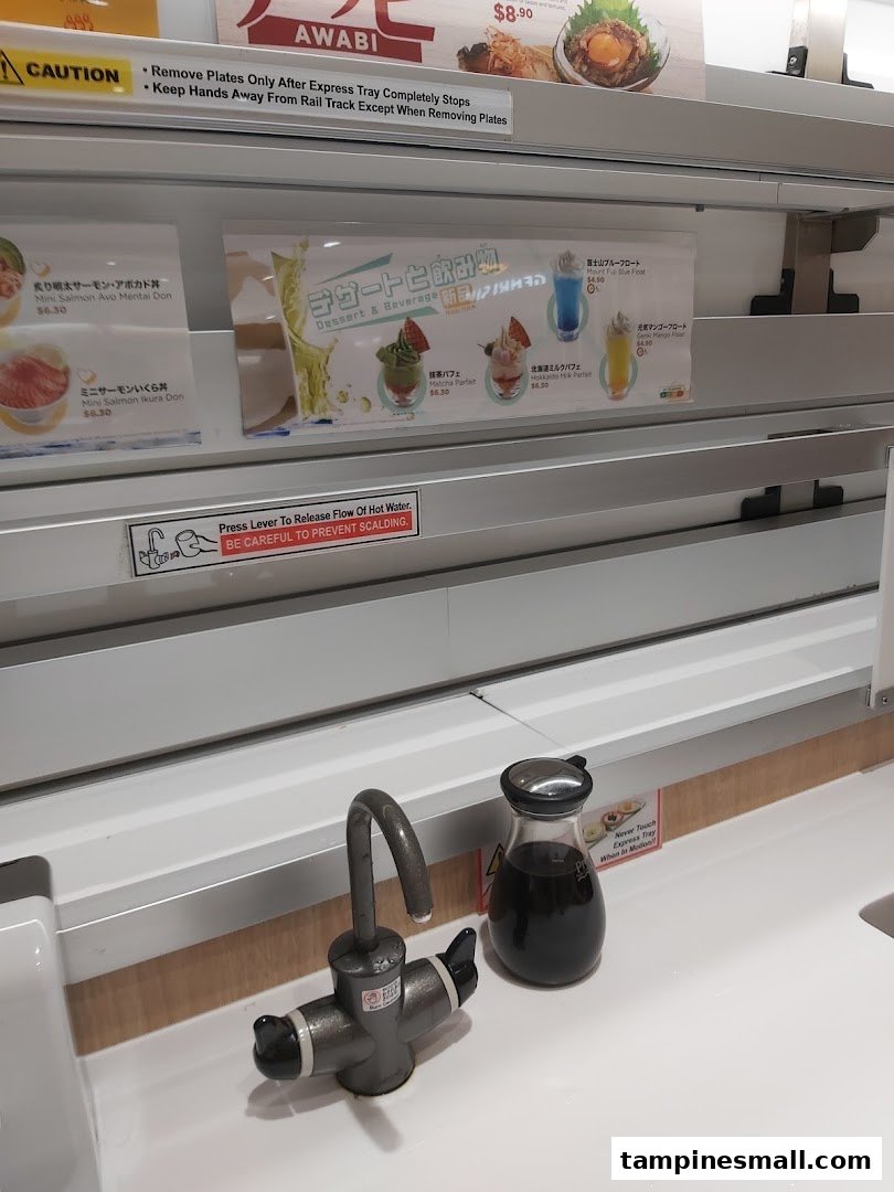 A close-up of a Genki Sushi dining counter with menu displays for desserts, mini-dons, and safety instructions.