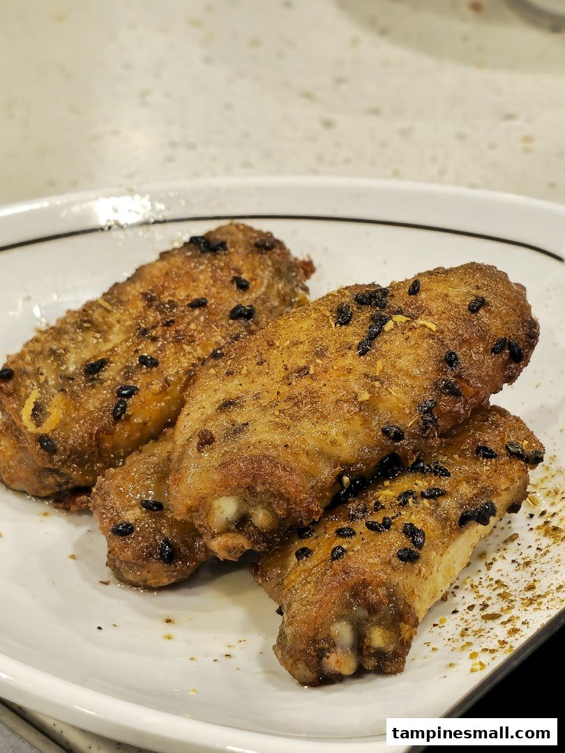 A close-up of four golden-brown seasoned chicken wings with black sesame seeds on a white plate.