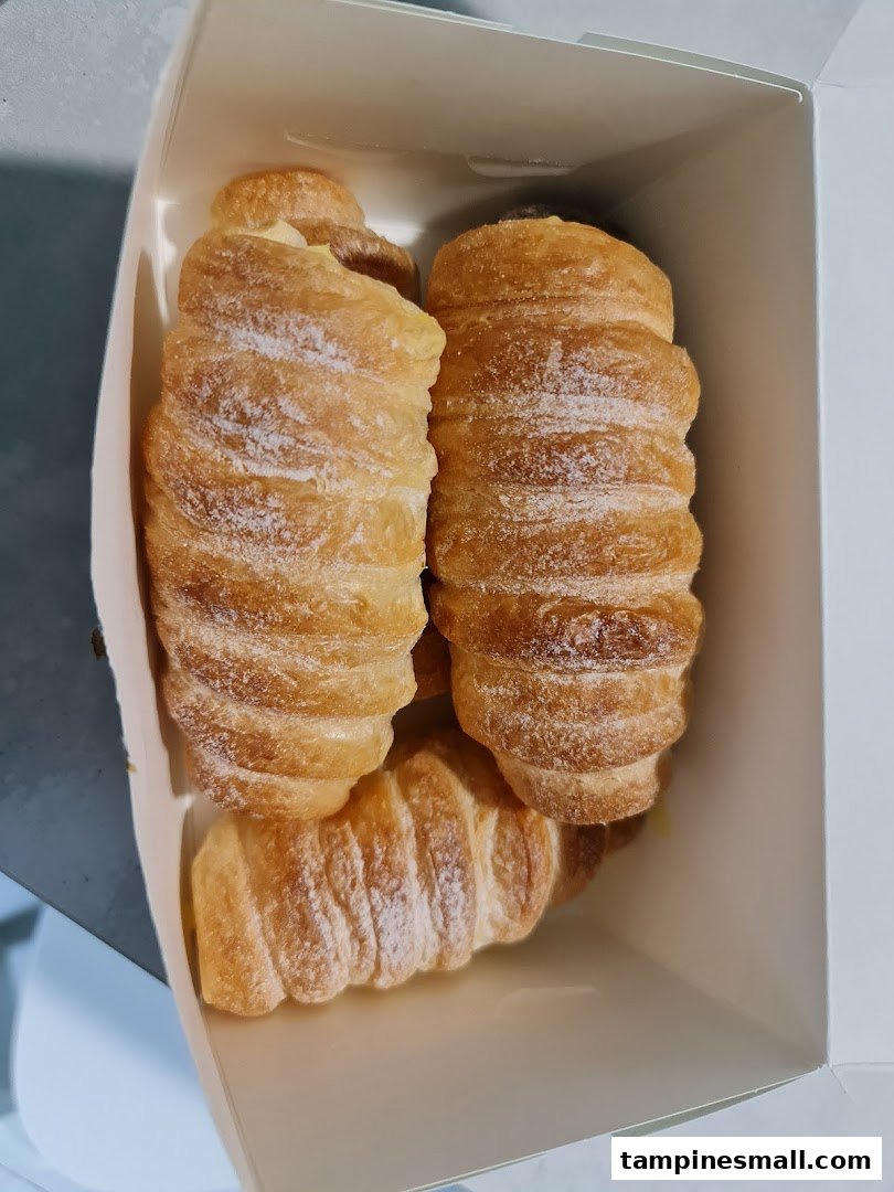 Three golden-brown pastries, possibly cream horns or croissants, dusted with powdered sugar in a white box.