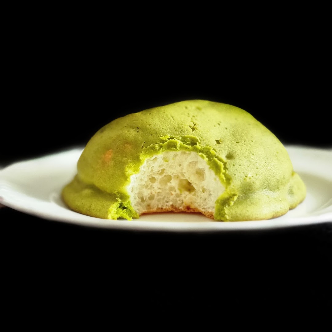 A vibrant green matcha melon pan, bitten to reveal its soft, fluffy interior, on a white plate.