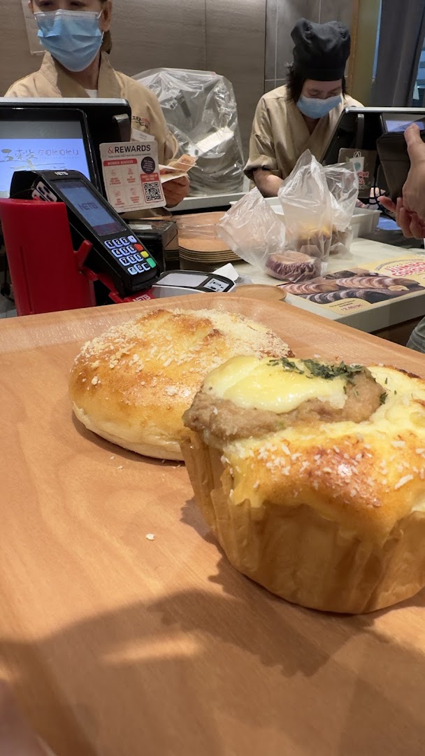 Freshly baked goods on a wooden tray at the Gokoku Japanese Bakery counter with staff in the background.