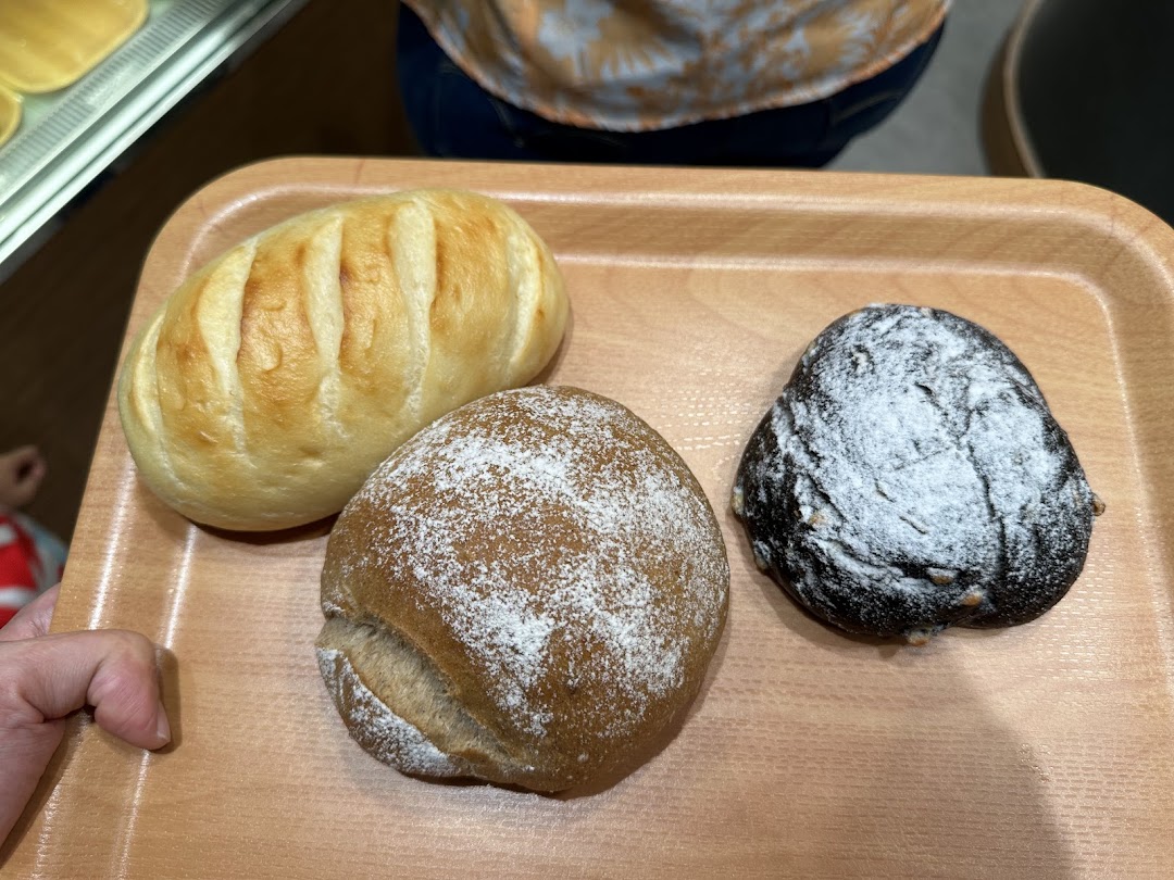 Three freshly baked Japanese breads, including a plain roll, a dark rye, and a chocolate bun, on a wooden tray.