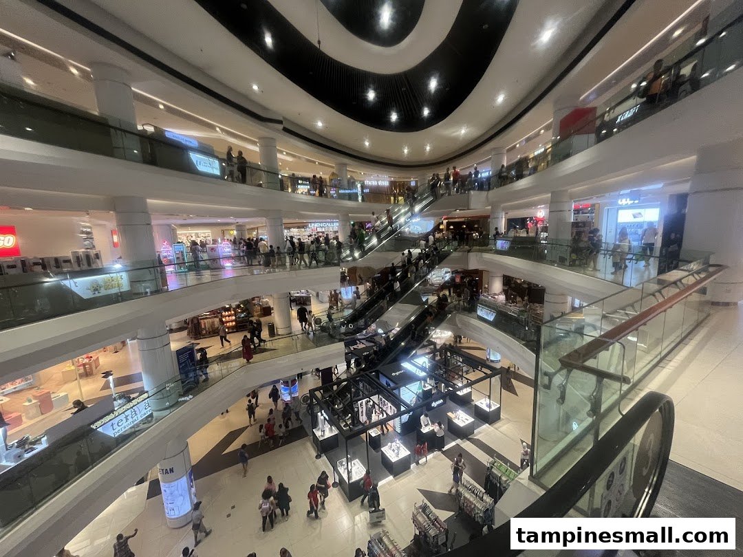 A bustling multi-level shopping mall interior with escalators, various shops, and many shoppers.