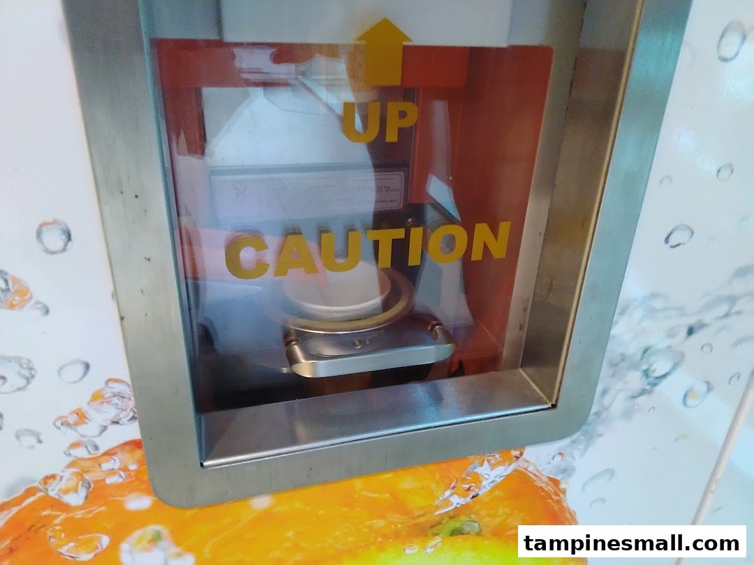 Close-up of an iJooz fresh juice vending machine's dispensing area with 'UP' and 'CAUTION' labels.