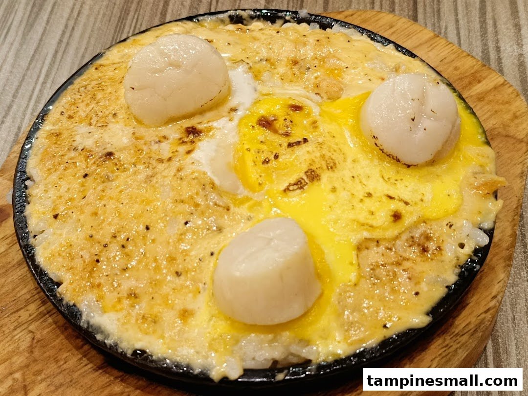 A sizzling hot plate dish featuring scallops, melted cheese, and a sunny-side-up egg.