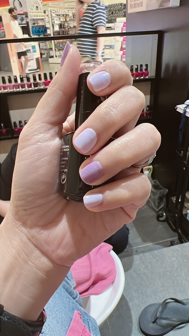 A hand showcasing fresh purple and lavender nail polish, holding a nail polish bottle inside a beauty salon.