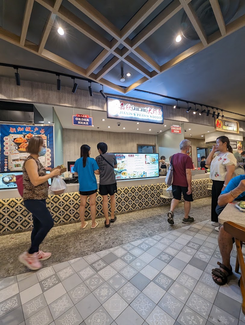 Customers queue and collect food at Jiaxin's Prawn Noodle stall in a bustling food court.