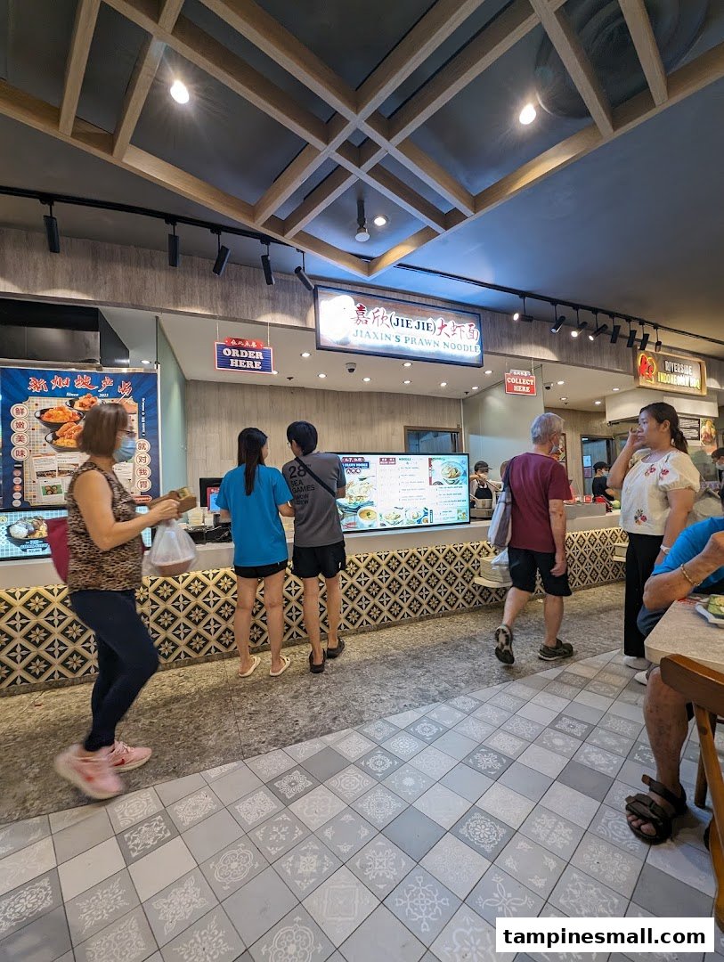 Customers queue and collect food at Jiaxin's Prawn Noodle stall in a bustling food court.