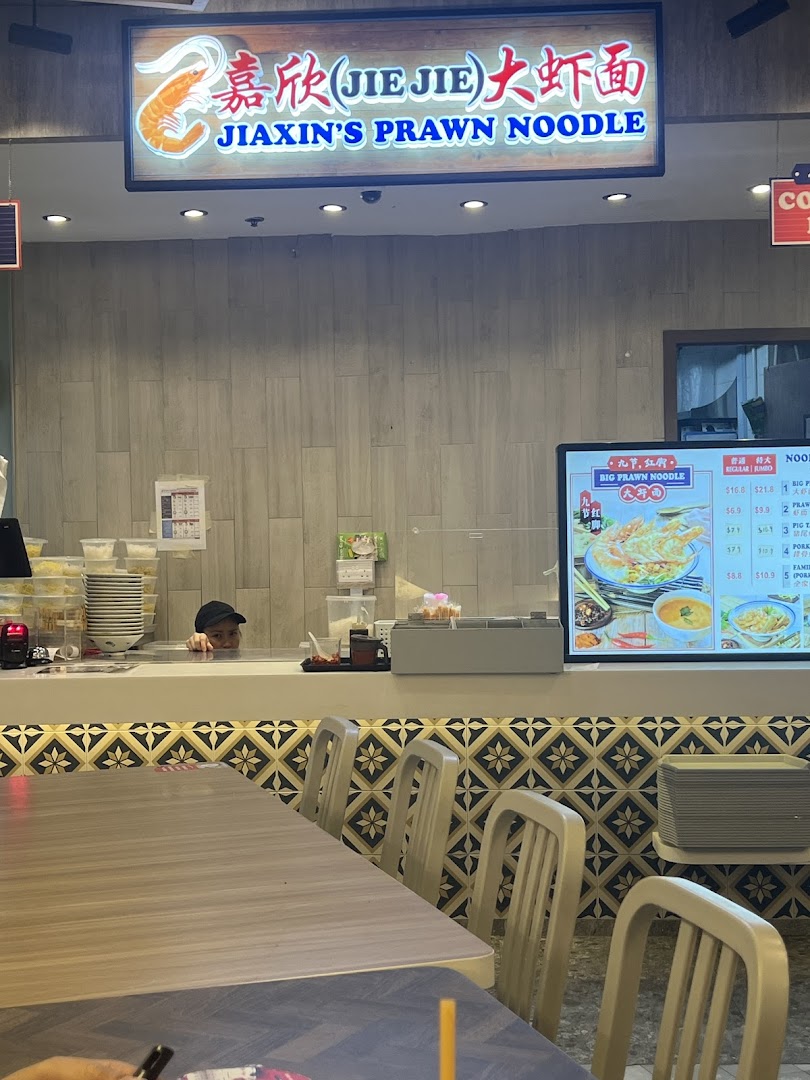 An interior view of Jiaxin's Prawn Noodle shop with a staff member and dining area.