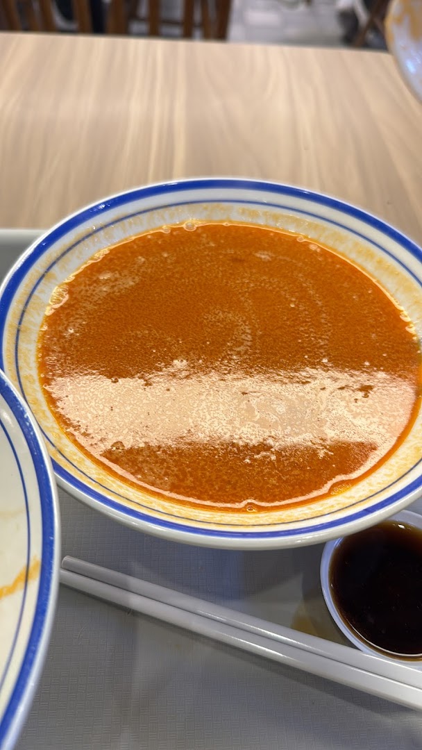 A close-up of a bowl of rich, orange-red prawn noodle soup with chopsticks and sauce.