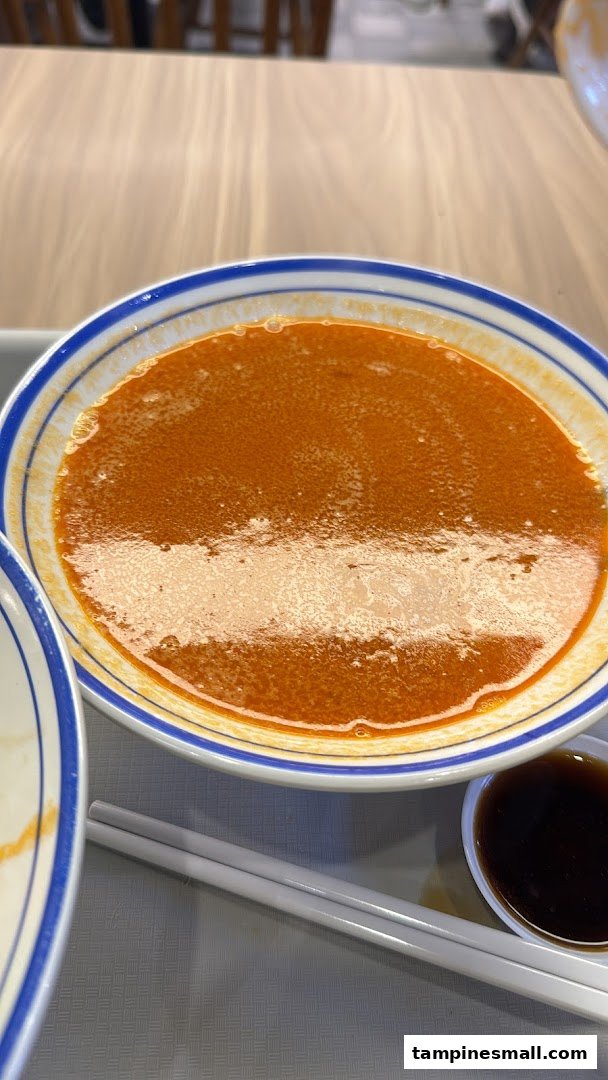 A close-up of a bowl of rich, orange-red prawn noodle soup with chopsticks and sauce.