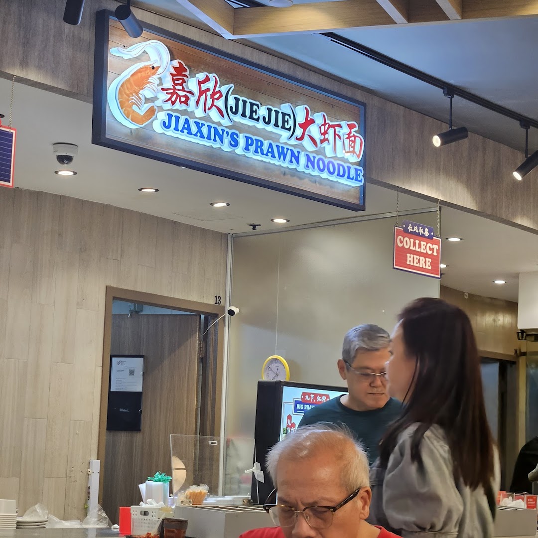 A view of Jiaxin's Prawn Noodle shop sign and service counter with customers.