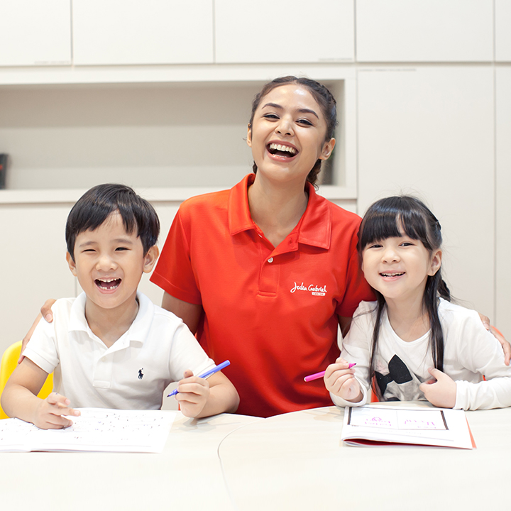 A smiling teacher engages happily with two young children during a learning session at Julia Gabriel Centre.