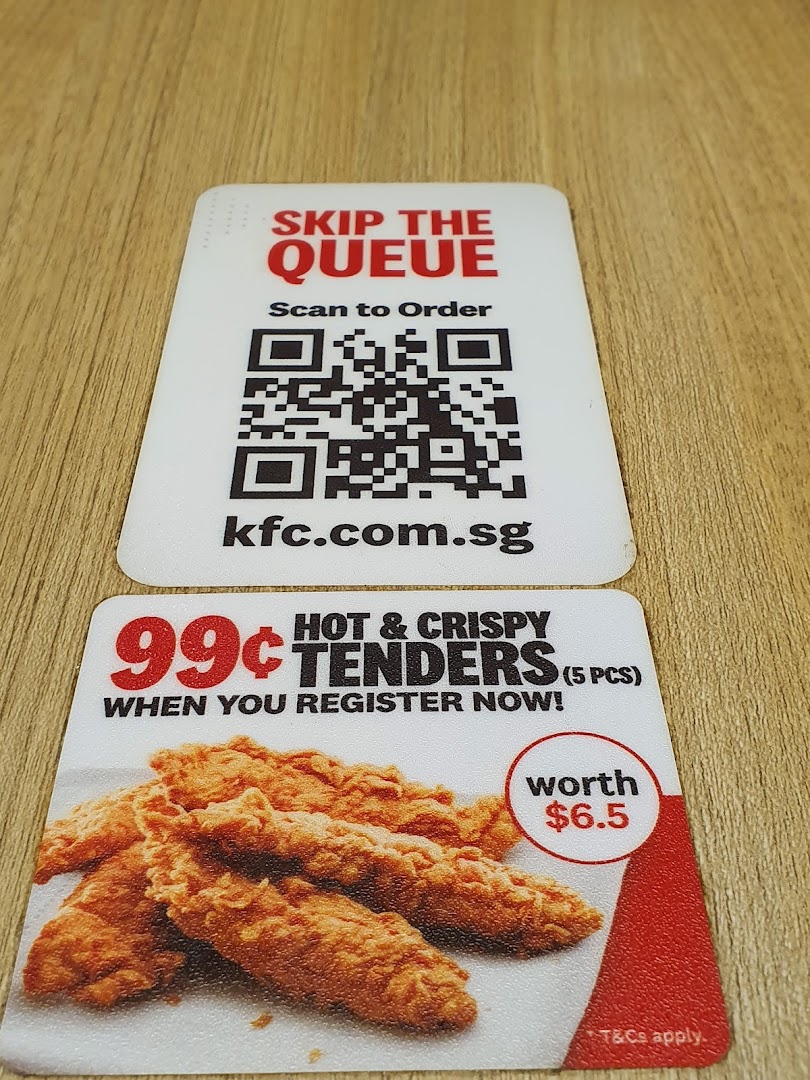 Two KFC promotional cards on a wooden surface, featuring a QR code for ordering and a chicken tenders offer.