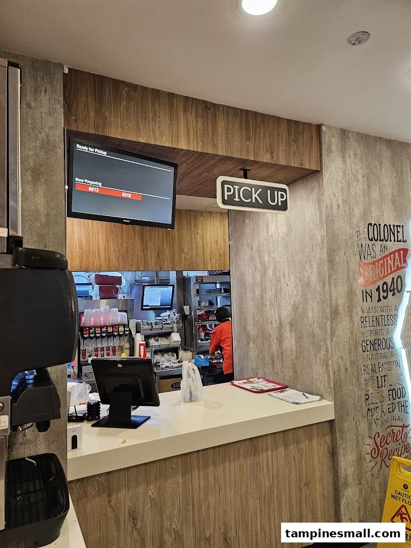 Interior view of KFC's pickup counter with a digital order screen and visible kitchen area.